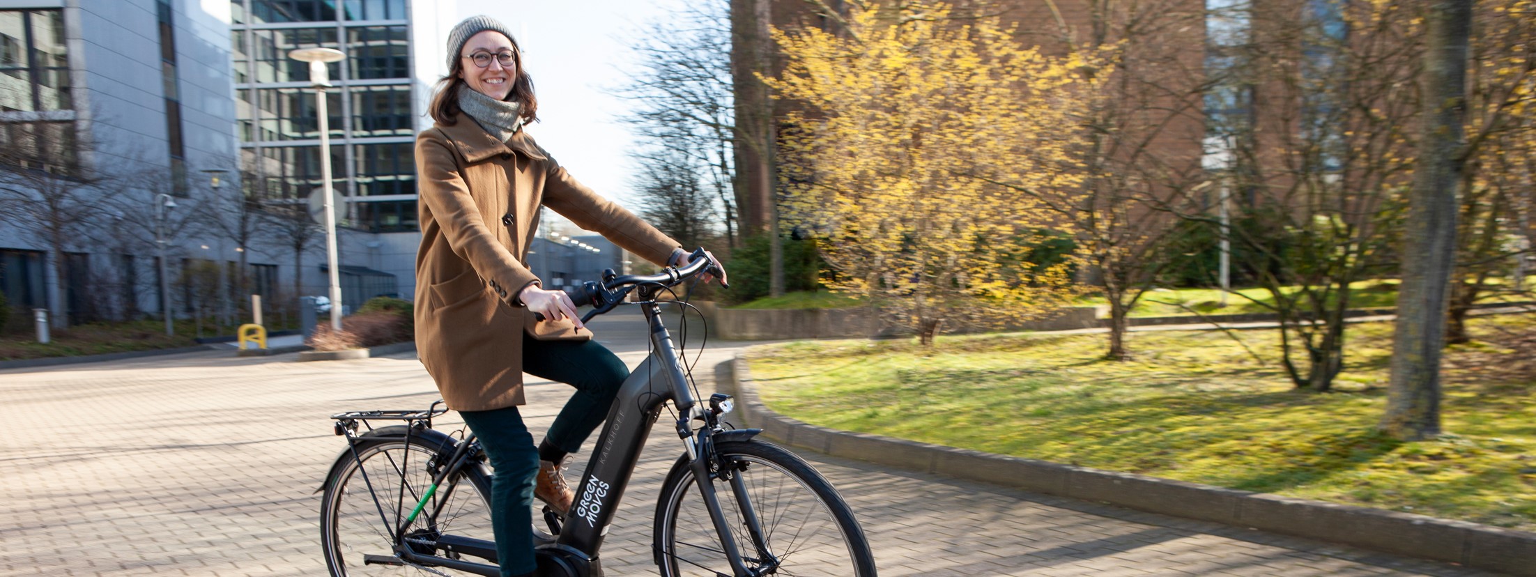 Eine Frau fährt ein Fahrrad auf einem Radweg. Sie trägt einen braunen Mantel, eine Mütze und eine Brille. Im Hintergrund sind Gebäude und Bäume zu sehen.