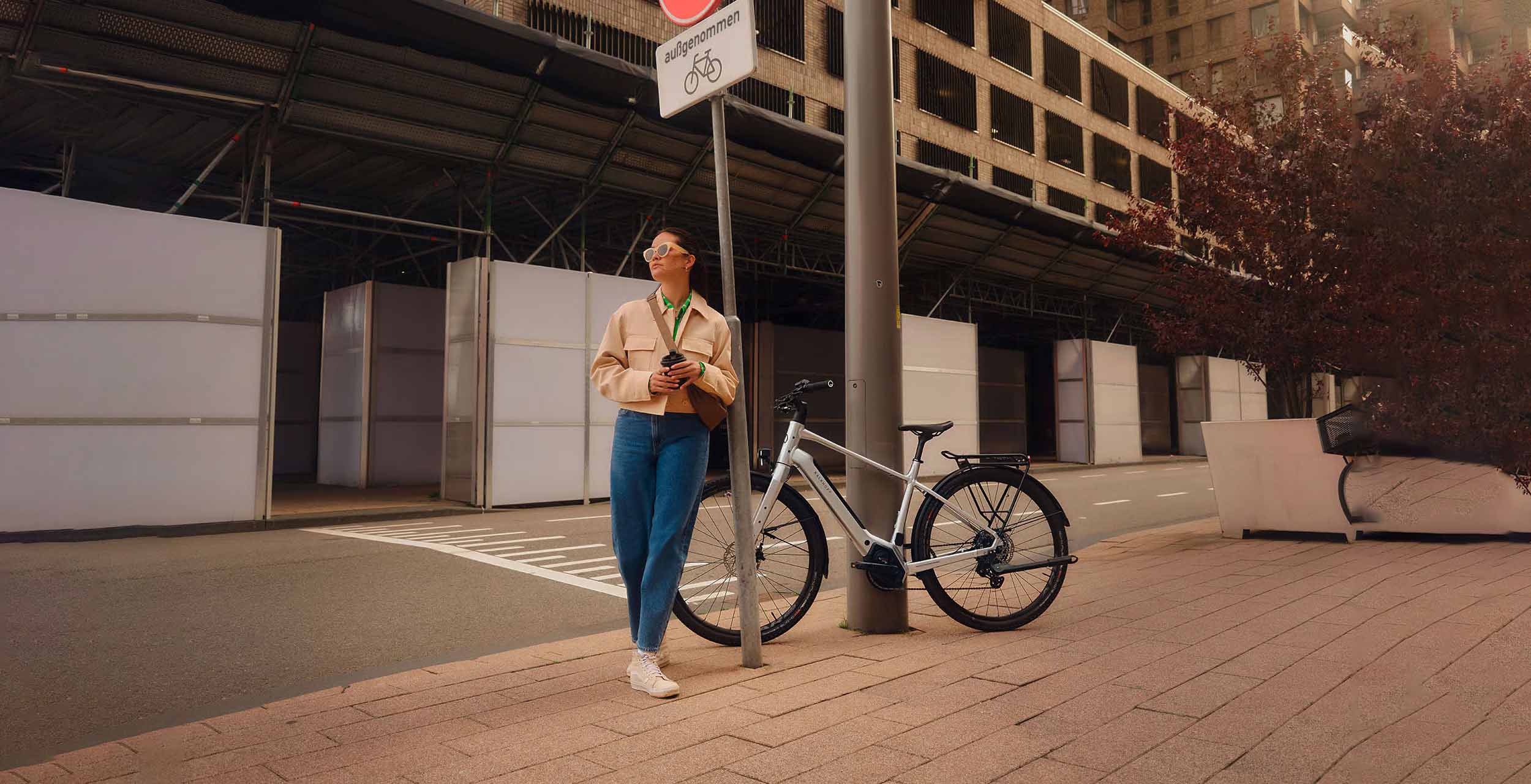 Junge Frau mit Sonnenbrille lehnt entspannt an einem Verkehrsschild, im Hintergrund steht ihr silbernes Rad der Marke "Kalkhoff".